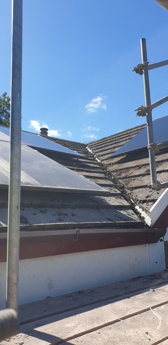 A shot of the gully where 2 sections of roof come together. There are 2 shiny new pv panels and the side of 2 of the older ones which are dull and dusty looking. There are scaffolding poles and the planks of a scaffold platform