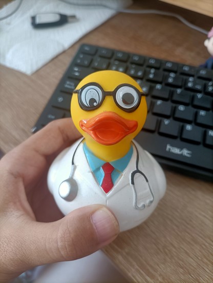 Una mano sosteniendo un pato de goma con un ojo más raro que el otro, disfrazado de médico. De fondo se aprecia el mismo teclado de antes