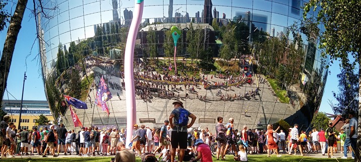 Landscape aspect. A crowd in front of a building, watching a bike race. But! The building is shaped like a drum and clad in a mirror-like covering, in which is reflected the race course (which is not otherwise visible to the viewer), and in the background, the skyscrapers and other buildings of central Rotterdam. 