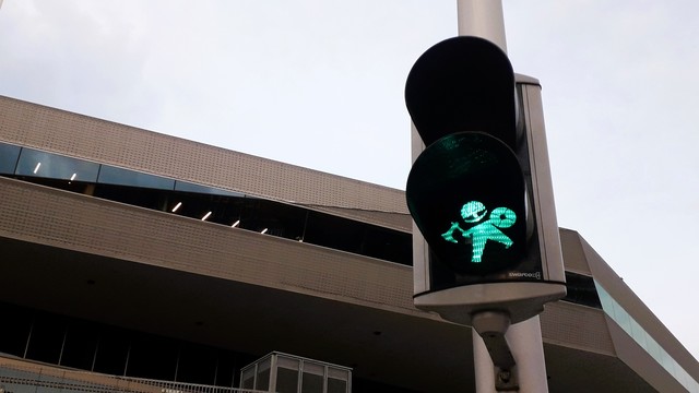 2019. Aarhus city centre, the roof of the library and white cloudy sky showing behind, looking up at the city’s first Viking pedestrian traffic light. It is showing the green ‘Walk’ figure.  Photo taken shortly after they were installed by Aarhus council. The 2019 Viking is sort of cartoonishly stumpy, in a cheery way. Their eyes show below their helmet. No horns, of course. They are at a jaunty, cocked angle. They’ve no mouth, but somehow still seem to be smiling. They’re swinging their battleaxe ahead and swinging their shield behind as if skipping happily across the road.