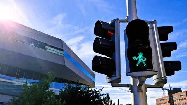 2024. Aarhus city centre, the roof of the library and blue sunny sky showing behind, looking up at the same Viking pedestrian traffic light as in the 2019 photo in this toot. The green ‘Walk’ figure is showing. Unlike the cheery 2019 Viking, the 2024 Viking looks deadly serious. They’re walking carefully erect and upright, battleaxe held in front of them, shield protectively close to their body. They have no eyes. Or mouth. They look expressionlessly cross and jaded, tired of this job.