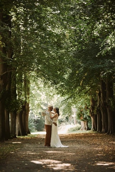 Bjarne and spouse, embracing in the park, shadowed by the foliage of the surrounding trees. Bjarne is wearing an Italian hat, a cream-colored shirt and copper colored pants. She is wearing a classic wedding dress, tailor made. We are facing each other, learning into a kiss.