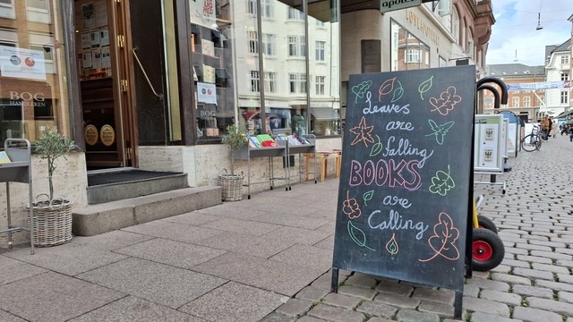 Chalkboard on the main square in Aarhus, outside Kristian F Møller independent bookshop. We see the cobble-stoned pedestrian street, people and bikes beyond. Books are displayed inside and outside the shop entrance. The staff have drawn lovely colourful autumn leaves around the edges of the blackboard and written in artistic and colourful lettering in the middle: 'Leaves are falling. Books are calling.'