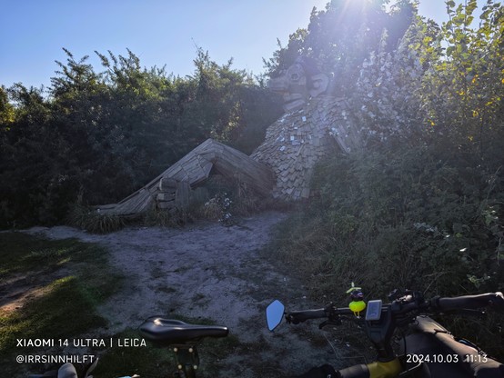 Ein Troll aus Holzteilen gebaut sitzt bequem am Strand zwischen bunten Büschen.