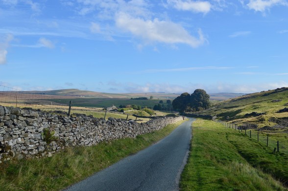 A road leads away into a wide shallow valley, edged on one side by an old stone wall with wooden fences every couple of yards, and on the other by grass falling into rough fields. In the distance, a small cluster of trees and farm buildings. The sky is an autumnal blue - fresh and clear. Patches of sunshine brighter like spotlights.