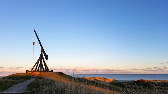 In the dunes in Skagen, a small fishing and seaside town on the literal tip of Jylland, mainland Denmark. It’s famous for its beautiful light. It is sunset, mid-summer, the White Nights when the sun barely dips below the horizon so it remains light all night. The sky is pale blue, cloudless except for a slim line of sunset-pink clouds at the bottom, as if bordering the horizon. Ahead, rolling dunes and marsh grass glow with green and gold colours. By contrast, the foreground is in shadow. Beyond, calm blue sea.  In front of us, a narrow gravel path leading up a slight incline to Vippefyret. The ‘tipping lantern’. It’s an exact reproduction of the original that was erected in 1627. It's an almost dramatic black wooden structure that rises about 10 metres high, like a tripod. A long boom juts out with a big metal bucket dangling on the end of it. Fire in the bucket served as a navigational point for sea vessels.