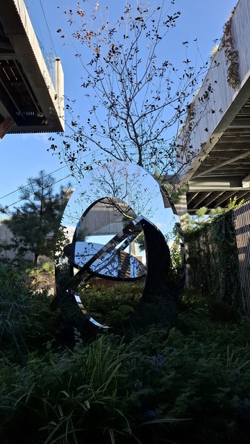 We are in an almost hidden corner of the public gardens on top of Salling department store on the main shopping street in Aarhus, 30 metres above the city. In front of us, a sculpture by Jeppe Hein called ‘The sky, the earth and you.’ It is framed below by lush green grasses and bushes in shadow; to the left and right by wooden fences and trees; and above by the underside of two wooden walkways. Behind it, a tall thin tree with thin branches dotted sparsely with small leaves. Above, a slice of clear bright cold-blue sky.  The sculpture comprises three nested circular mirrors that tilt in different directions, reflecting fragments of the surroundings. The effect is rather magical, I think. From this angle, the top of the largest mirrored ring reflects the thin tree and the sky in a way that makes it look as if the reflection is an actual part of the tree. The sky and trees and bushes and shadows and light blend in the rings to make a whole new landscape.