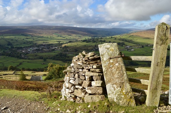 Sunlight on an old stone gate post. Beyond, views down into a wide valley criss-crossed with walled fields. Brown swathes of autumn bracken cutting through the green, and in the base of a valley, a cluster of houses looking tiny due to the distance. The sky is partly blue, clouds casting large shadows - a sense of freshness and openness and space.