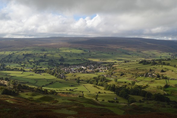 View down into the base of a valley from an elevated point. There is autumn sunlight breaking through clouds and illuminating a network of green walled fields. In the centre, a small town is clustered round an open market place. The sun is catching on a row of white houses. Beyond, open moorland over low hills.