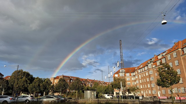 Frederiksbjerg, mid-city Aarhus, a road with cars in the foreground. Beyond: tall mature trees and lovely old redbrick apartment blocks. The sky is dark blue with clouds with that peculiar lightfall that you get when the weather has been switching between strong cold sunshine and torrential rain showers. Curving in from the right, a double rainbow gleams through the sky.