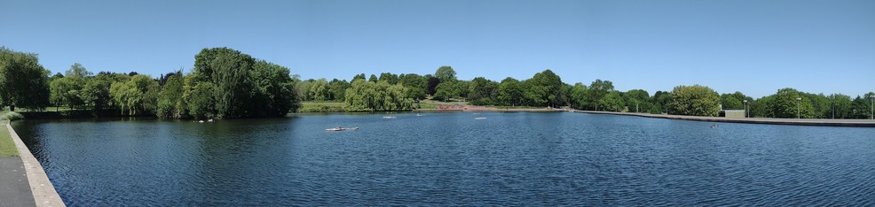 Panorama of the lake at Dartmouth Park, Sandwell, West Midlands, England. Clear blue sky with water below separated by a line of trees across the horizon.