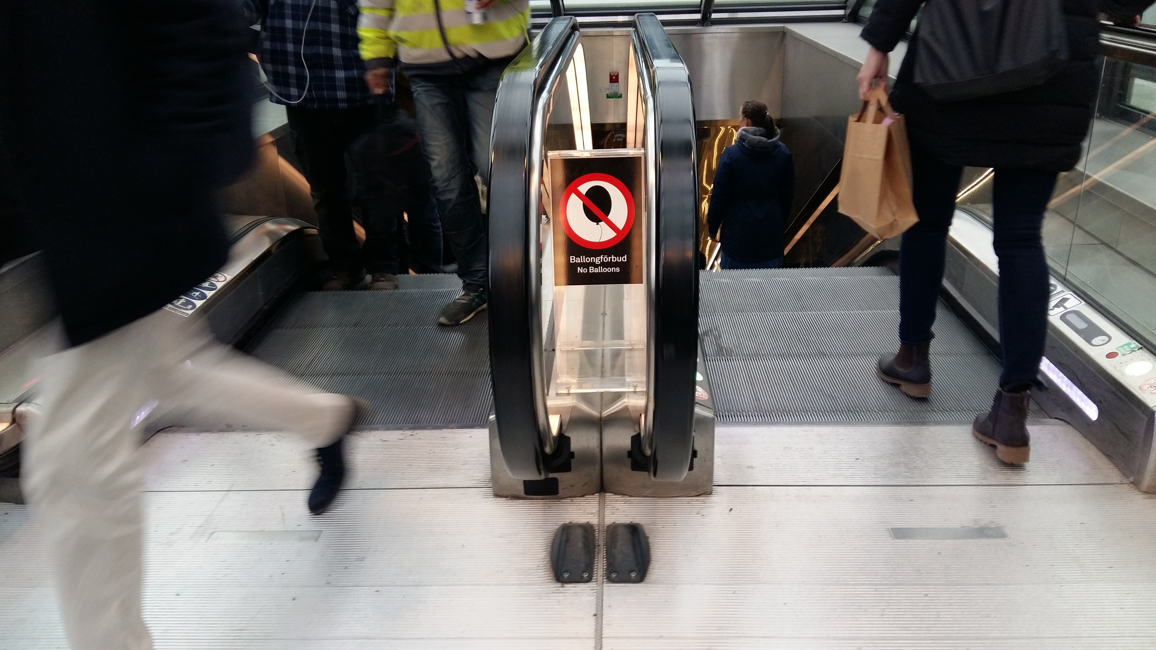 Close-up of top of an escalator at Malmö railway station, Sweden. We see the lower legs of people getting off and on, some blurry in motion, in mid-step. There is a space in the middle, between the Up and Down handrails. In it hangs a sign. Black background. Centred, a white circle bordered in red with a line through it - the This is Forbidden sign. Inside it, the forbidden item that it is crossing out: A party balloon. It is black, above white text that says in Swedish and English Ballongforbud / No balloons.  
