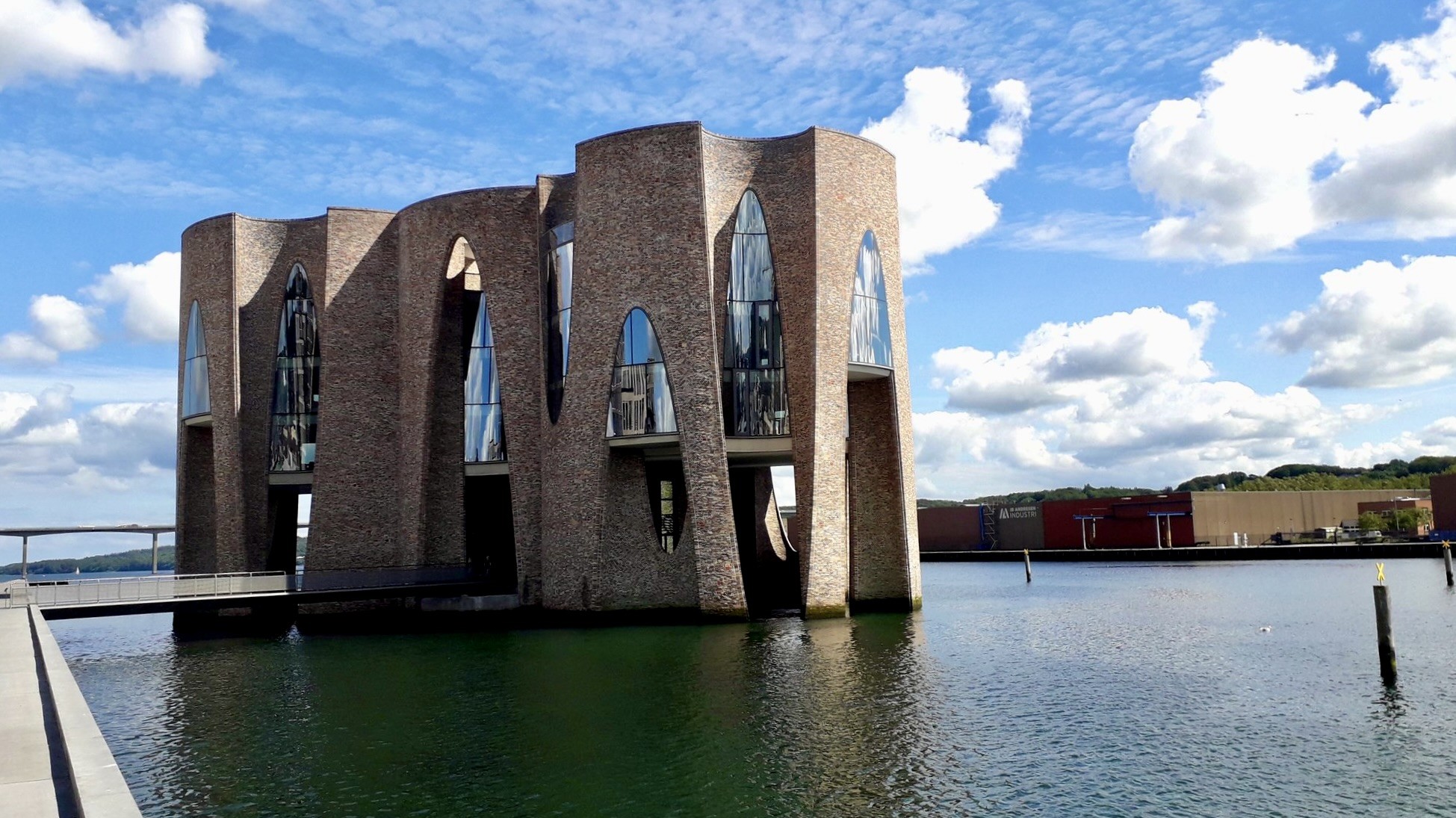 Short version: A brick structure with cylindrical sections and mirrors, built in a fjord. Fuller description: Seen from a slight distance, across a short wooden bridge, as if floating in the water, Olafur Eliasson's architectural artwork Fjordenhus – the house on the fjord. It is on the water in Vejle Fjord, Denmark. Commissioned by Vejle Kunstmuseum (Art Museum), it comprises four interconnected cylinders with a height of 28 meters. The brickwork and angles are gorgeous, both seen together and individually. The overall structure, seen here, is both bombastic and inviting at the same time. It sort of suggests waves or sails on the water, but at the same time has a slight look of a fortress.