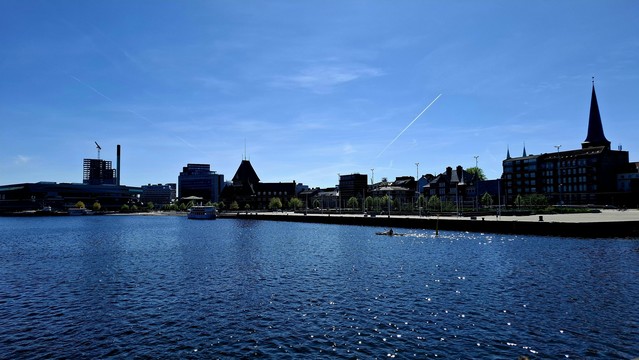 Short version: Aarhus harbour and city skyline. Fuller description: the harbour and coast road skyline including turrets and spires. A hot Summer day. The sky is light blue. The sea is deep blue, covered in silver sparkles from the sunlight. Small in the distance, a sightseeing boat. Closer, a single kayak. A white trail from a plane cuts diagonally through the sky, like a daytime comet. The scene is very blue and all is calm. 