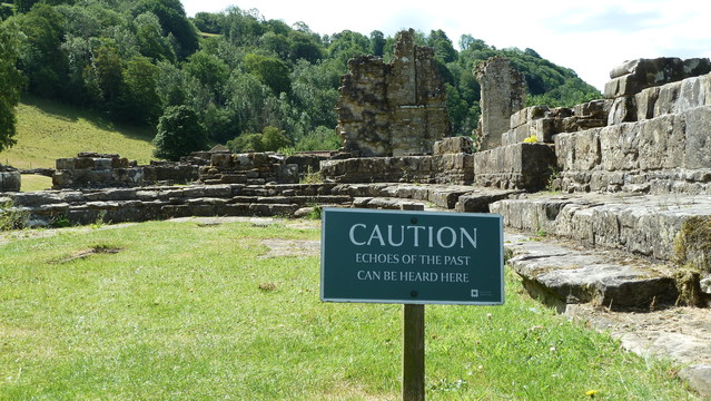 Stone remains of a Medieval abbey standing in grassland with trees in the background. Green metal sign in the foreground reads: CAUTION Echos of the past can be heard here 