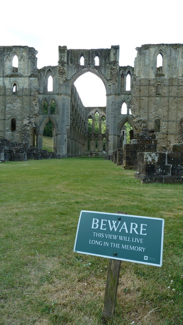 A ruined Medieval abbey set against a grey sky. In the foreground a green metal sign standing in a large grassy lawn.