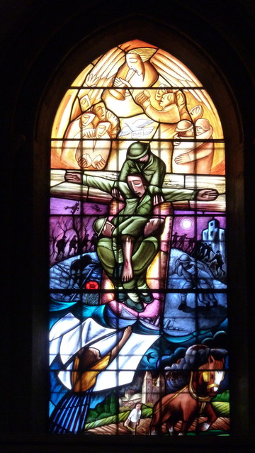 Stained glass window. In the centre three soldiers are seen one lifting the other under the arms, one below under his legs. The outstretched arms are symbolic of the cross. The design fuses Christian iconography with familiar Great War imagery to reiterate the horrors of the 1914-18 conflict whilst emphasising the need for forgiveness and reconcilliation.