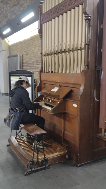A small pipe organ located in the undercroft of a Victorian railway station. You player seated wearing a white baseball cap and black clothes.