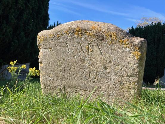 A stone grave marker engraved "AMV 1797". The stone is set in grass, trees in the background and a blue sky.