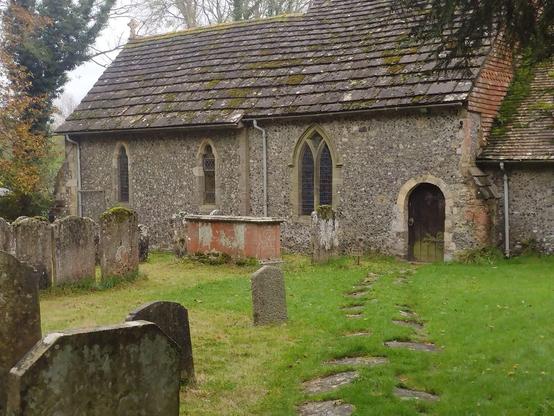 A church in a grassy churchyard. A row of stepping stones lead to a small door with around arch on the right of the building. The door is closed.