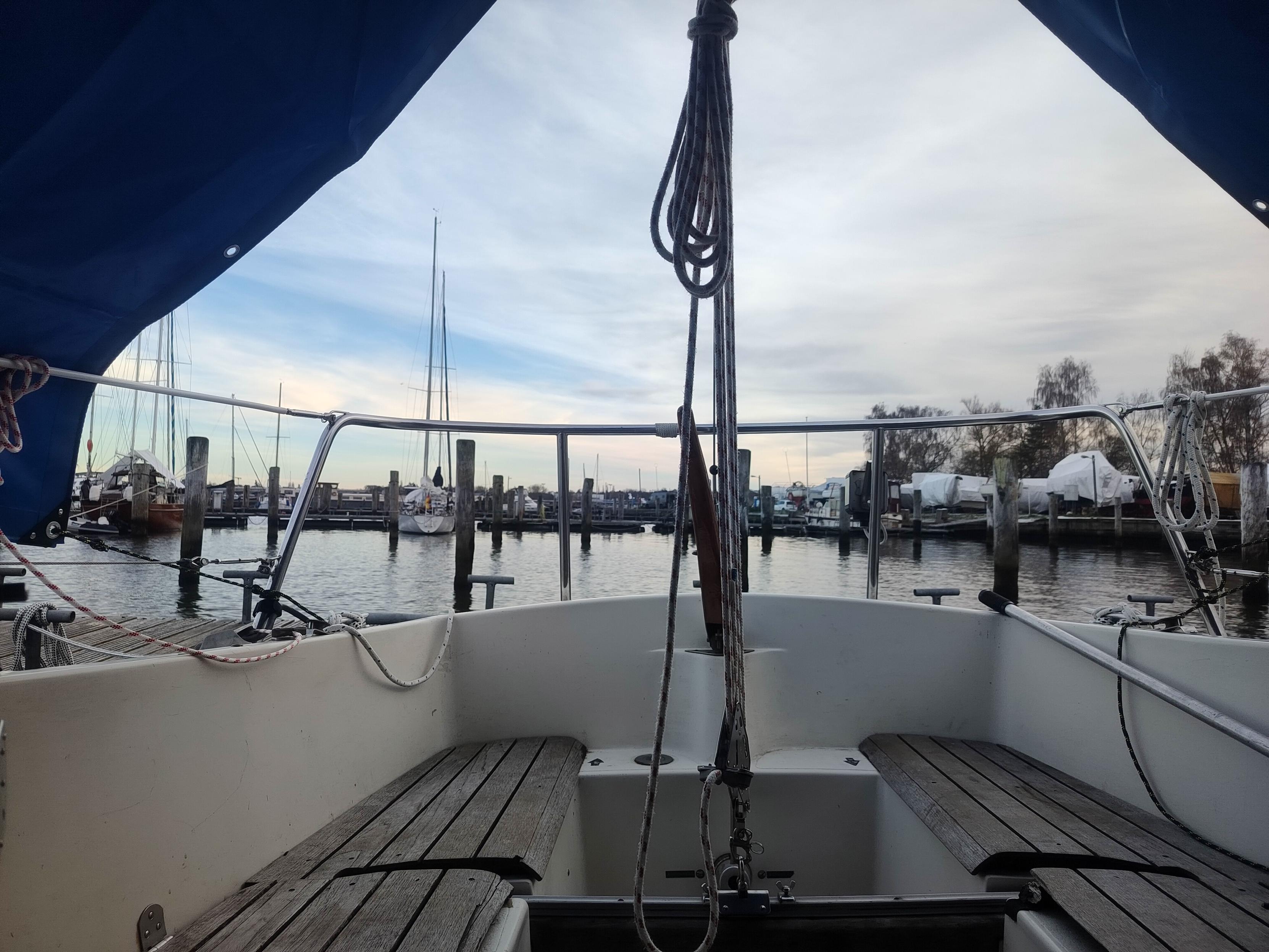 Ein Bild aus der Sicht einer Person, die auf einem Boot sitzt und auf das Wasser blickt. Das Boot hat einen weißen Innenraum und ein blaues Dach. In der Mitte des Bildes befindet sich ein schwarzer Mast. Das Wasser ist ruhig und es gibt viele Boote im Hintergrund, die an einem Dock festgemacht sind. Der Himmel ist bewölkt und es sind Bäume in der Ferne zu sehen.
