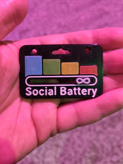 A hand holding a black tag indicating the ‘social battery’ of the bearer. There are four bars from high (blue) to low (red). Below the bars is a movable indicator. It is almost at the minimum.