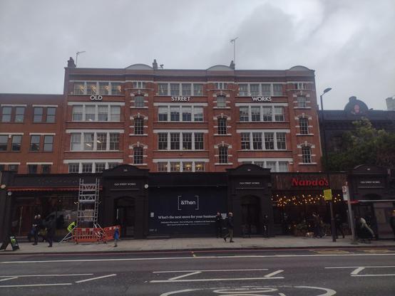 An Edwardian five story commercial and office building. Red brick, flat roof. White windows on the upper floors, black fronted retail units at street level. Grey sky above.