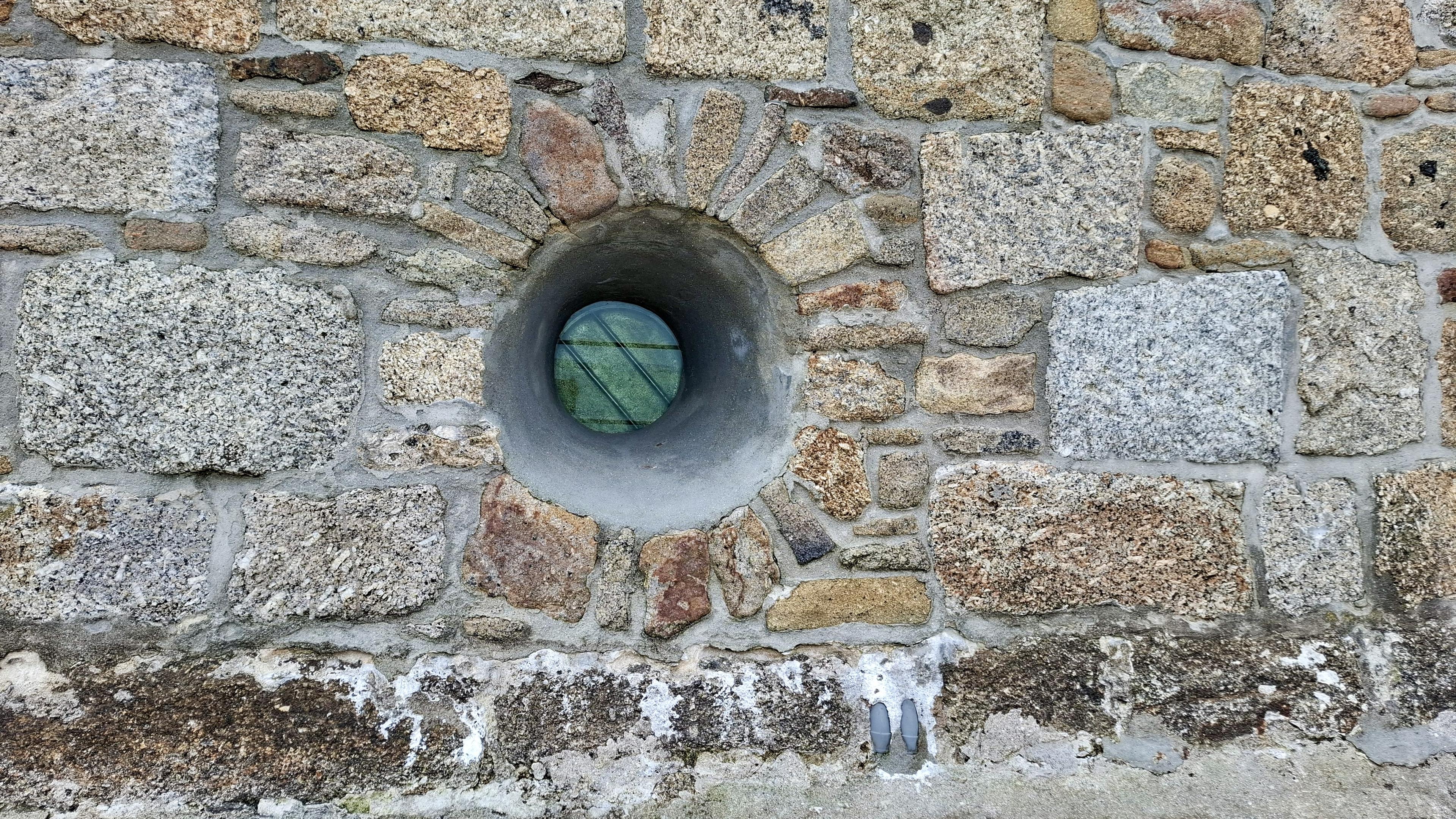 Close-up of a picturesque old wall with grey and beige bricks. Recessed deeply in it, a tiny little round window, barely bigger than an eye. It is light green glass. This is a random wall by a harbour in Cornwall. I don't know what the window is for or what's behind it and am happy not to know.