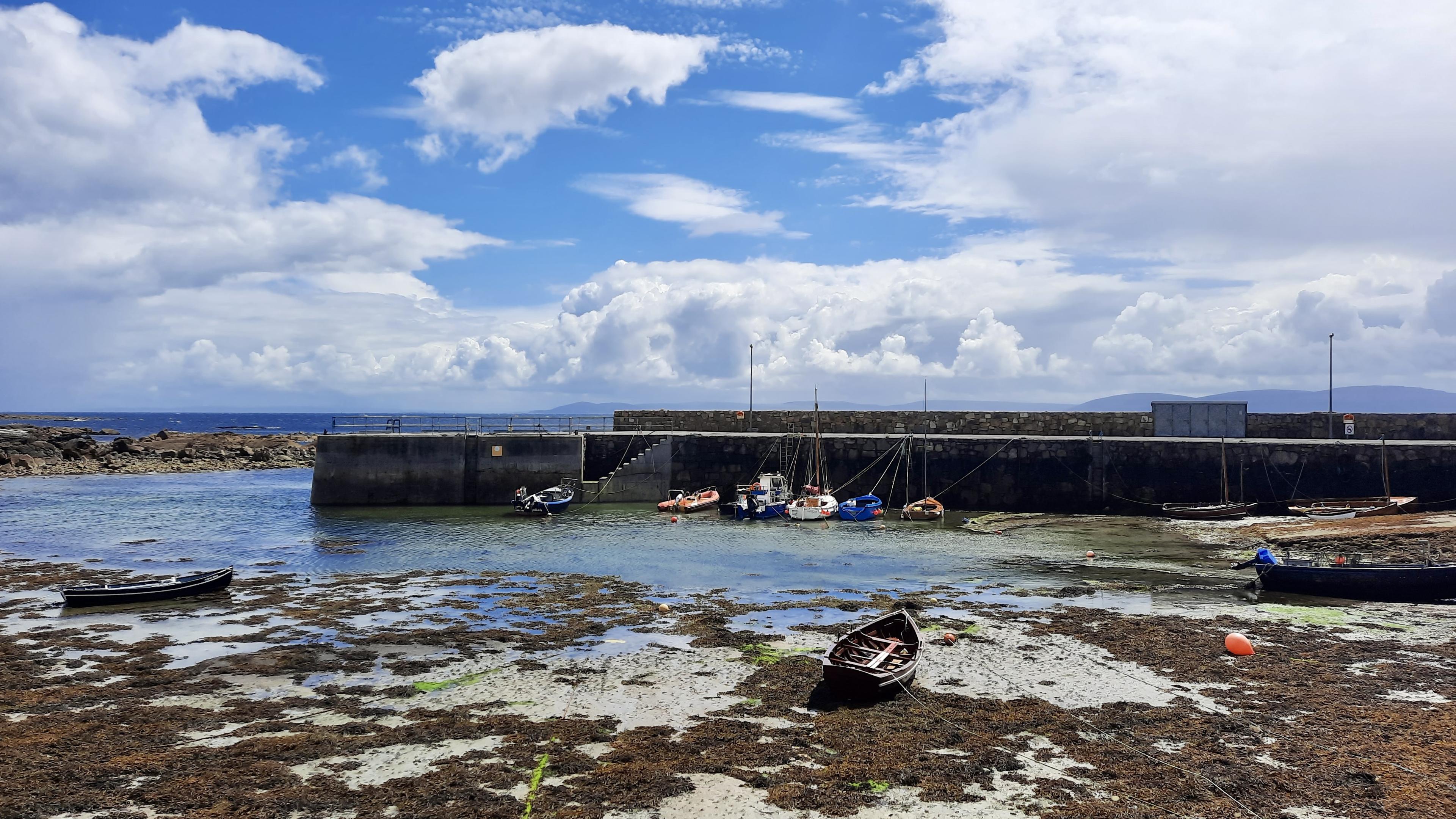 Short version: a harbour at low tide. Fuller description: Looking from a slight height down at a little local harbour in An Spidéal - Spiddal. Galway. Ireland. A dark grey stone pier wall creates a sheltered inner harbour. The tide is out. Small boats are scattered about, beached on light brown sand and thick dark brown seaweed. Beyond, a glimpse of open sea, the Atlantic Ocean, which was in disguise that day because it is light bright sunny blue. Above it, an equally bright blue sky with huge bulging white clouds, so low and big that they almost look like cartoon clouds.