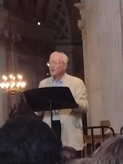 Diarmaid MacCulloch speaking to an audience at St Paul's Cathedral, London. Standing behind a lectern, pale stone pillar to the right and an illuminated chandelier to the left.