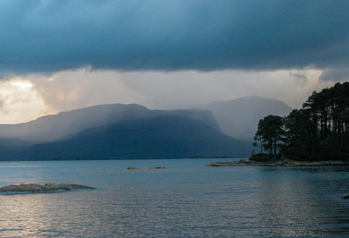 A  view across a loch to musty stormy mountains. A bit of s conifer-covered  island is in the foreground, on the right