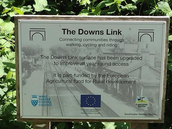 A sign near the site of the former #Henfield station, which reads (in part) The Downs Link, Connecting communities through walking, cycling and riding. The Downs Link surface has been upgraded to improve all year round access. It is part-funded by the European Agricultural fund for Rural development.

