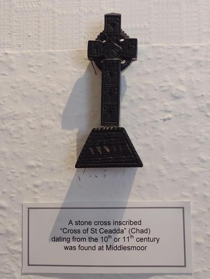 A dark brown cross mounted on a white wall. Caption card below.