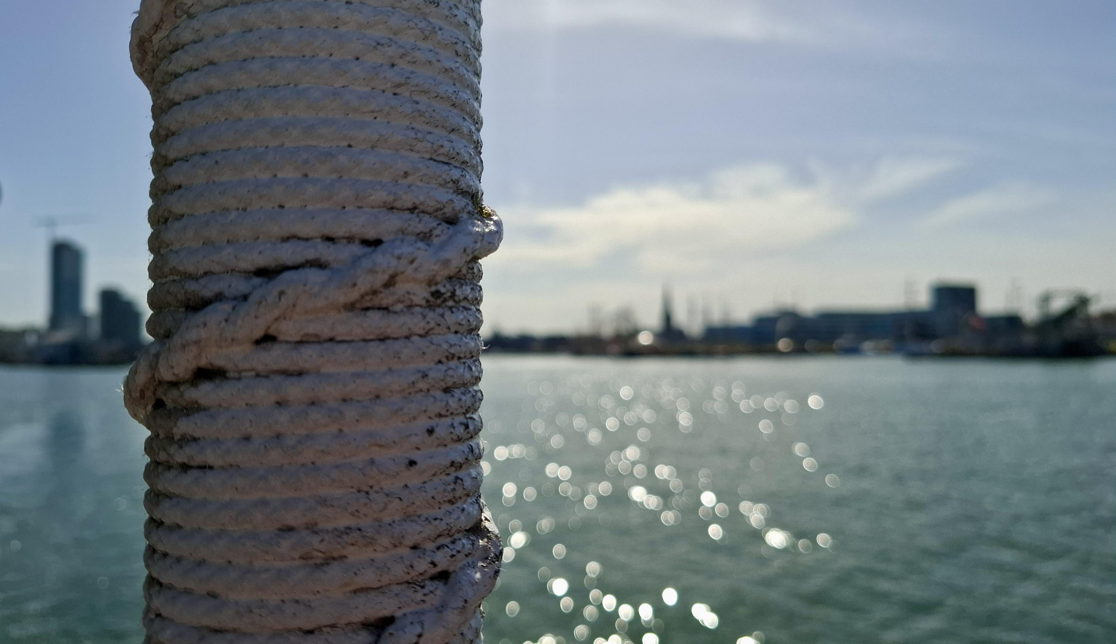 Short version: sharp foreground, blurry background, sea and skyline. Fuller description: Foreground, to the left, close up, a worn metal white pole with metal rope entwined around it. Blurry in the distance, green sea water speckled with blurry dots of sunlight. Beyond, almost in silhouette from facing into the high sun, a wavelength of the Aarhus city skyline - buildings and spires in different heights.