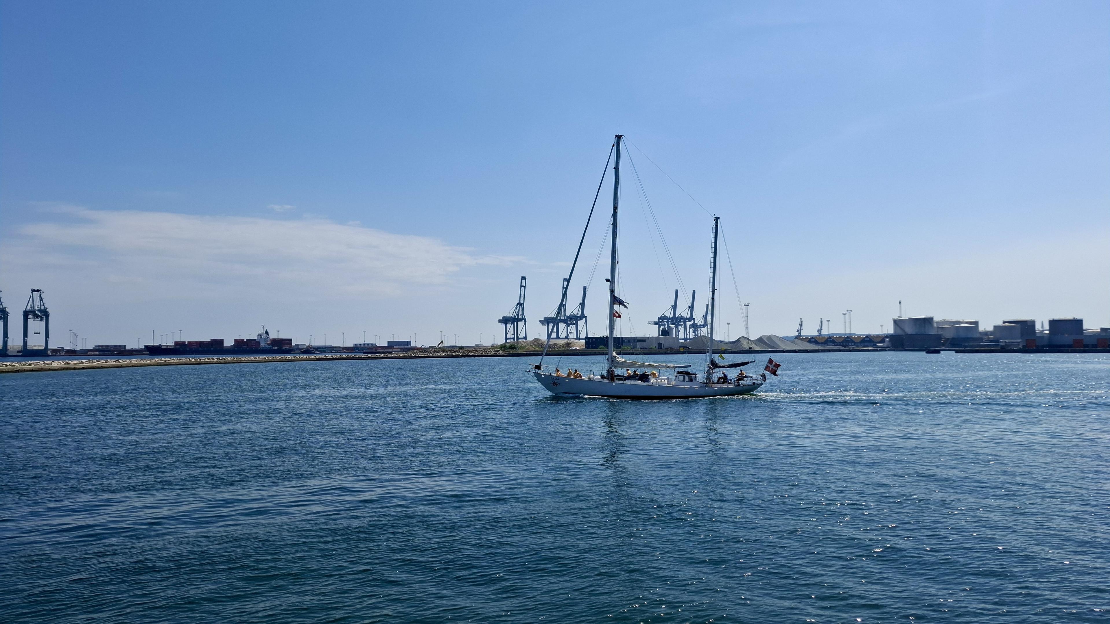 Short version: the ship sailing. Fuller description: deep blue sea, almost cloudless scorching-hot blue sky. Near the Aarhus harbour entrance. The Sail-out Parade, the Tall Ships Race 2026 leaving Aarhus, Denmark, with no wind, so no sails up. Docks and cranes and port entrance opposite. Unseen – local boats and crafts filling the bay to the left, waving goodbye; the other Tall Ships sailing out, waiting on one another in the bay or lining up to sail out from harbourside themselves. The crew of the Tara - 18m hull and tallest mast 19.6 m – is lined up on deck, waving and shouting exchanges with the crowds where we are standing on the pier. The photos show the ship turning in a perfect circle, drawing an actual circle of different-coloured blue water on the surface of the sea.