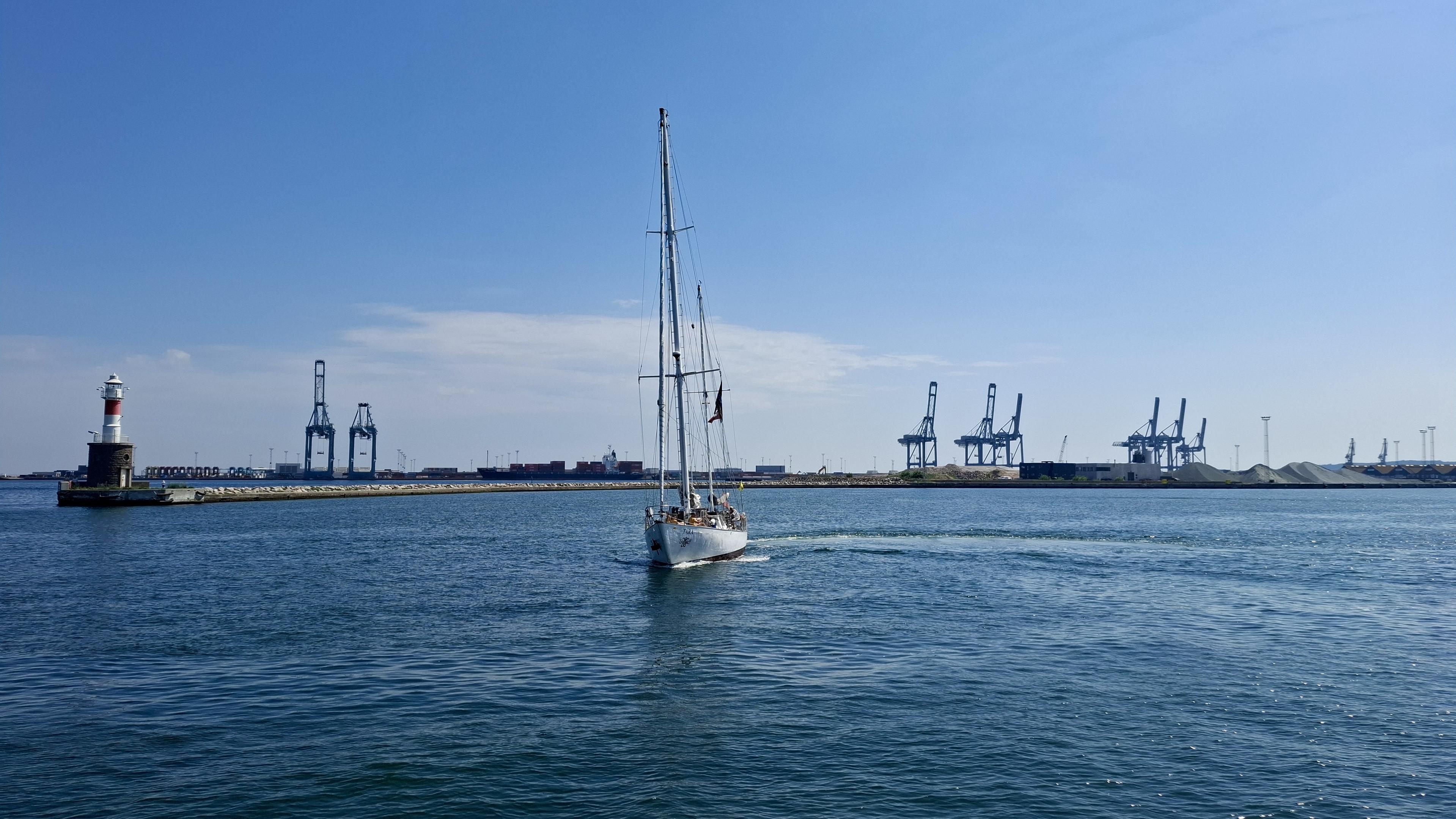 Same alt text as other photos. Short version: the ship sailing. Fuller description: deep blue sea, almost cloudless scorching-hot blue sky. Near the Aarhus harbour entrance. The Sail-out Parade, the Tall Ships Race 2026 leaving Aarhus, Denmark, with no wind, so no sails up. Docks and cranes and port entrance opposite. Unseen – local boats and crafts in the bay to the left, waving goodbye; the other Tall Ships sailing out, waiting on one another in the bay, or waiting to sail out from the harbourside themselves. The crew of the Tara - 18m hull and tallest mast 19.6 m – is lined up waving and shouting exchanges with the crowds on the pier. The photos show the ship turning in a perfect circle, drawing an actual circle of different-coloured blue water on the surface of the sea.