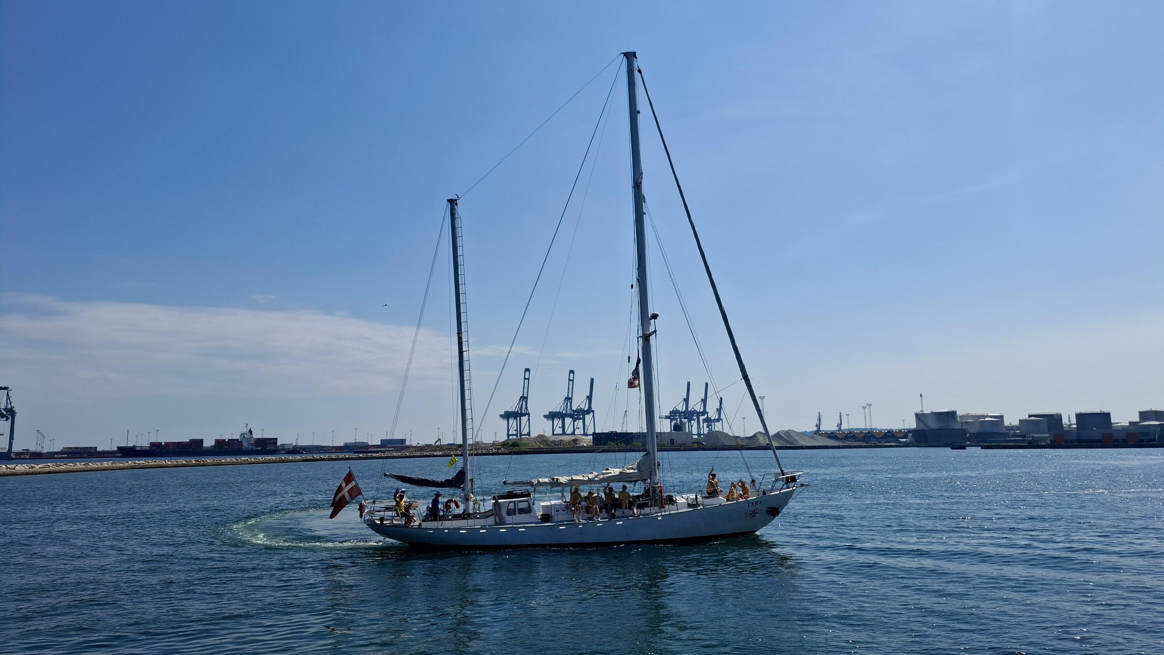 Same alt text as other photos. Short version: the ship sailing. Fuller description: deep blue sea, almost cloudless scorching-hot blue sky. Near the Aarhus harbour entrance. The Sail-out Parade, the Tall Ships Race 2026 leaving Aarhus, Denmark, with no wind, so no sails up. Docks and cranes and port entrance opposite. Unseen – local boats and crafts in the bay to the left, waving goodbye; the other Tall Ships sailing out, waiting on one another in the bay, or waiting to sail out from the harbourside themselves. The crew of the Tara - 18m hull and tallest mast 19.6 m – is lined up waving and shouting exchanges with the crowds on the pier. The photos show the ship turning in a perfect circle, drawing an actual circle of different-coloured blue water on the surface of the sea.