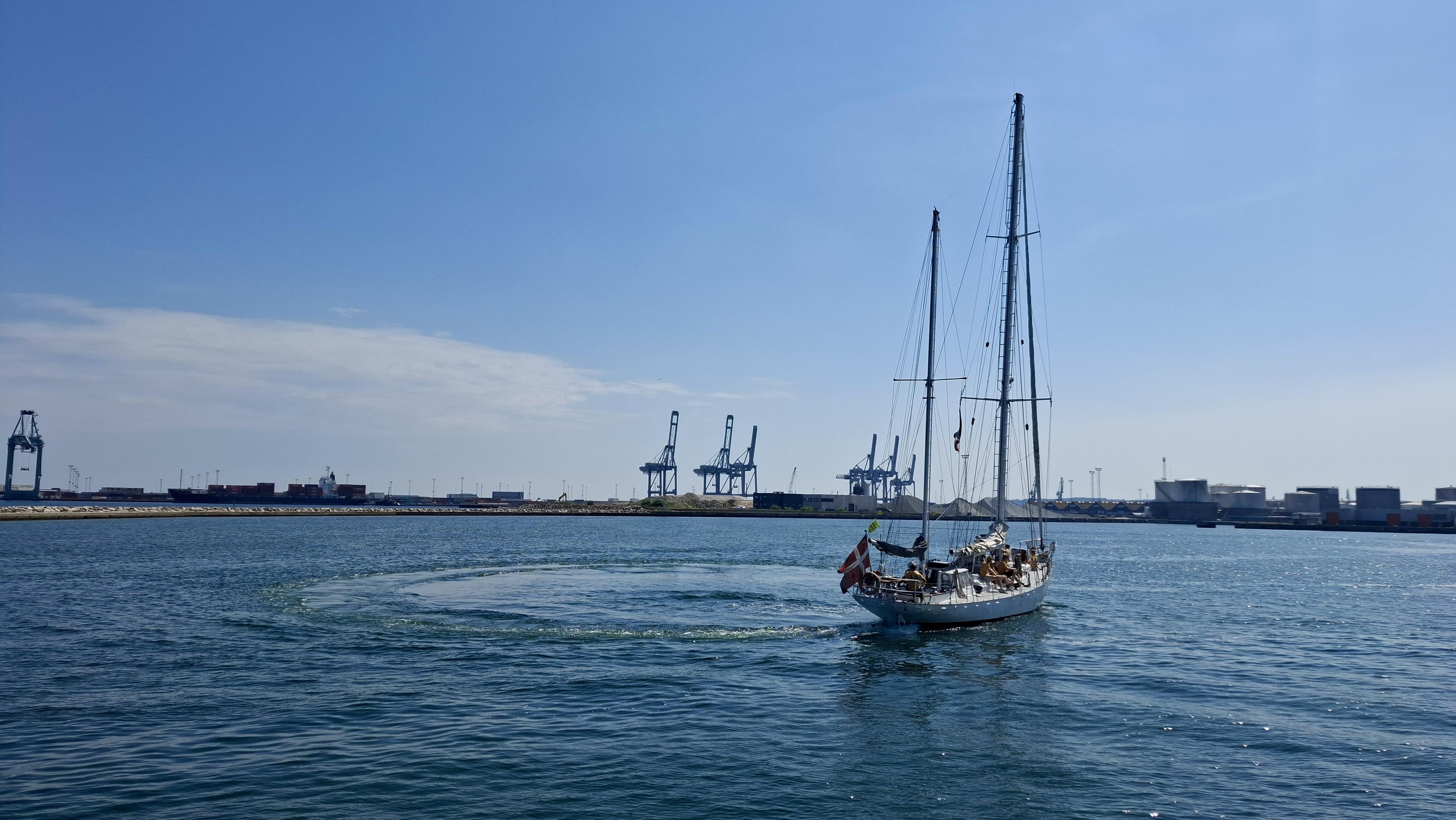 Same alt text as other photos. Short version: the ship sailing. Fuller description: deep blue sea, almost cloudless scorching-hot blue sky. Near the Aarhus harbour entrance. The Sail-out Parade, the Tall Ships Race 2026 leaving Aarhus, Denmark, with no wind, so no sails up. Docks and cranes and port entrance opposite. Unseen – local boats and crafts in the bay to the left, waving goodbye; the other Tall Ships sailing out, waiting on one another in the bay, or waiting to sail out from the harbourside themselves. The crew of the Tara - 18m hull and tallest mast 19.6 m – is lined up waving and shouting exchanges with the crowds on the pier. The photos show the ship turning in a perfect circle, drawing an actual circle of different-coloured blue water on the surface of the sea.