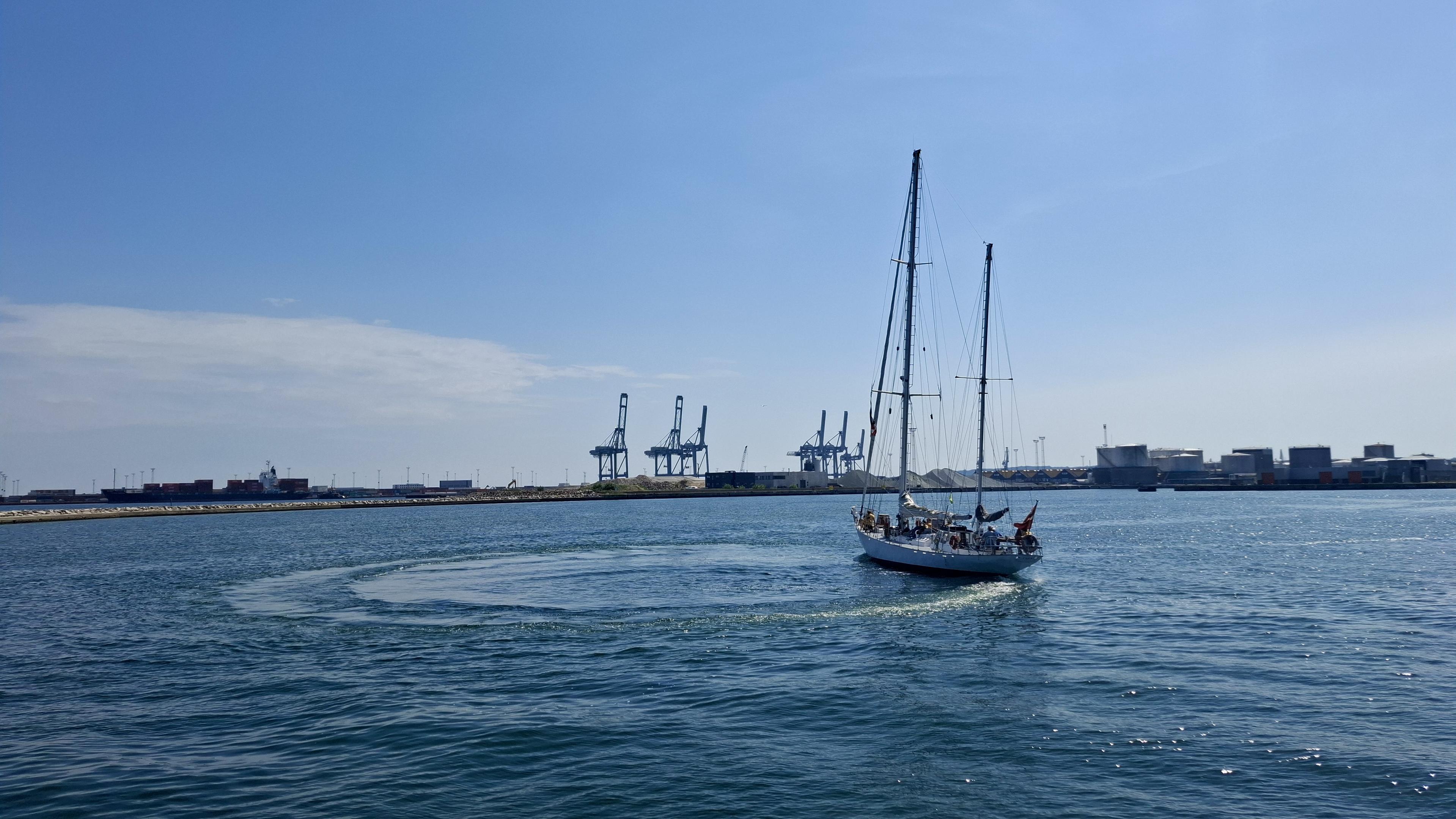 Same alt text as other photos. Short version: the ship sailing. Fuller description: deep blue sea, almost cloudless scorching-hot blue sky. Near the Aarhus harbour entrance. The Sail-out Parade, the Tall Ships Race 2026 leaving Aarhus, Denmark, with no wind, so no sails up. Docks and cranes and port entrance opposite. Unseen – local boats and crafts in the bay to the left, waving goodbye; the other Tall Ships sailing out, waiting on one another in the bay, or waiting to sail out from the harbourside themselves. The crew of the Tara - 18m hull and tallest mast 19.6 m – is lined up waving and shouting exchanges with the crowds on the pier. The photos show the ship turning in a perfect circle, drawing an actual circle of different-coloured blue water on the surface of the sea.