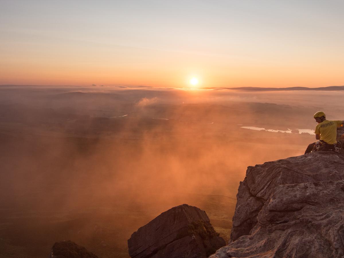The sun sets on a distant horizon whilst misty clouds form in the valley below. A  climber with helmet and t-shirt sits on the top of a crag at the far right, looking down over the scene.