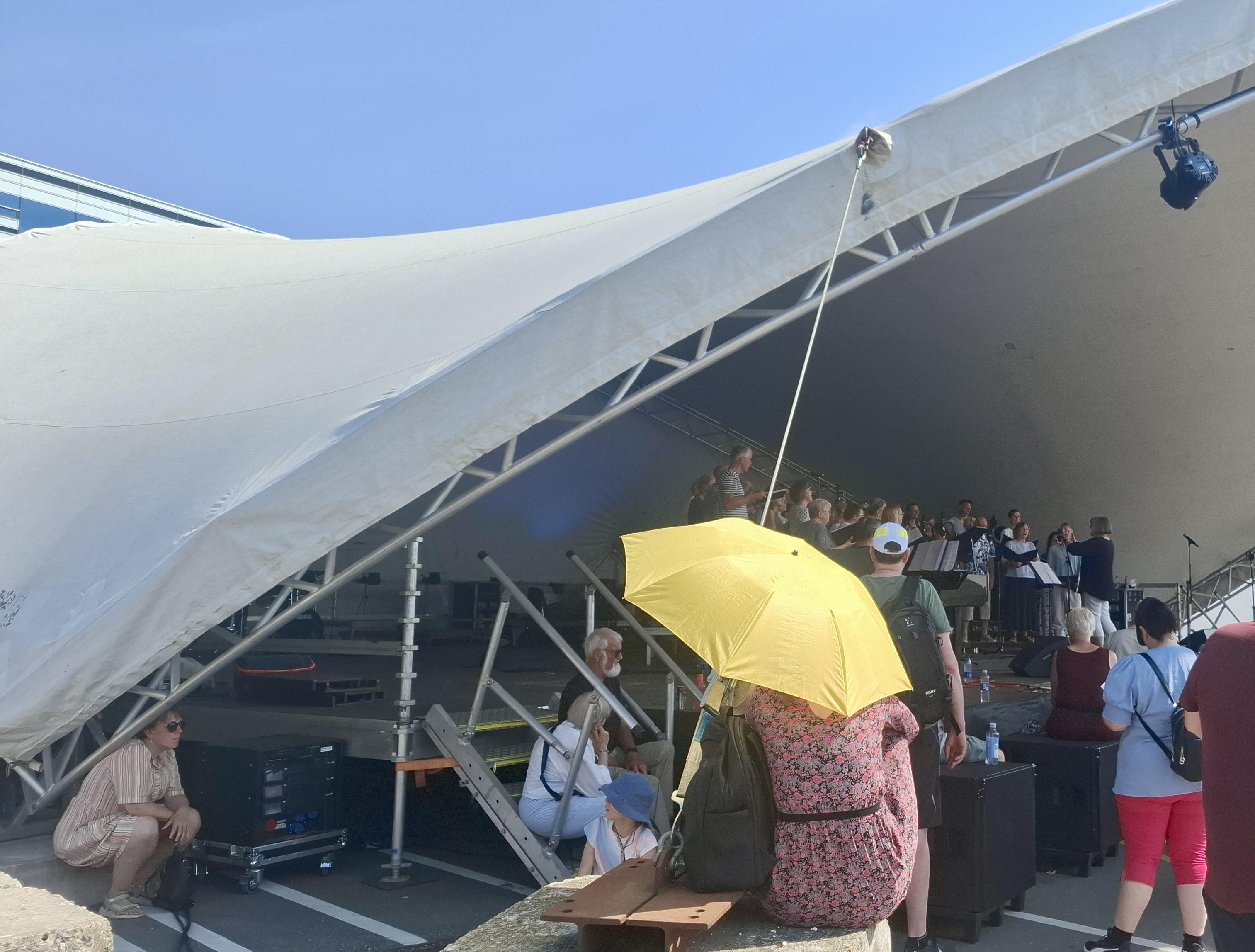 Small crowd near or under a marquee, cloudless blue sky above, a choir singing on stage. In front of us, a yellow umbrella being used by someone as a parasol