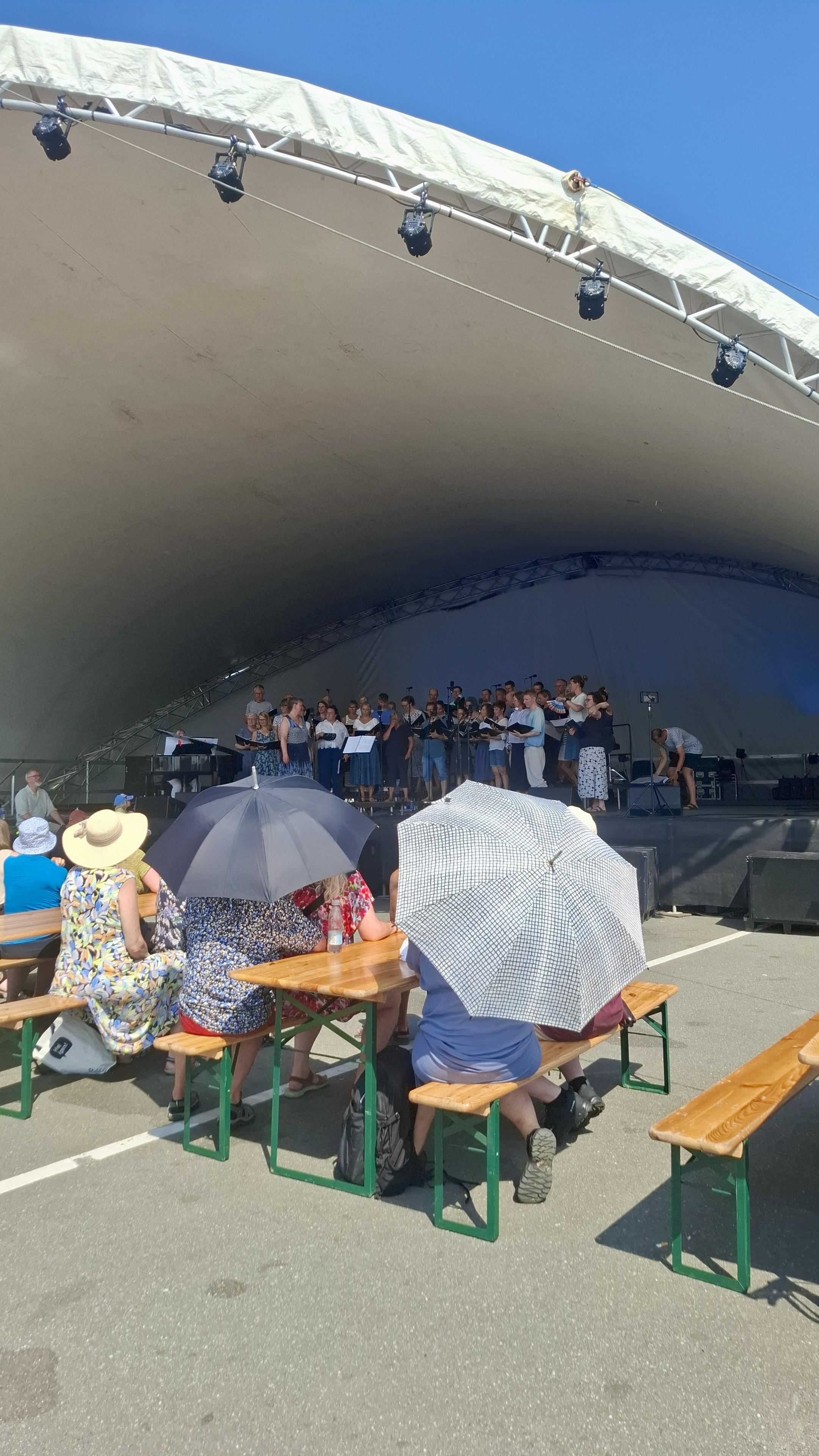 Small crowd near or under a marquee, cloudless blue sky above, a choir singing on stage. In front of us, people on benches using rain umbrellas as parasols