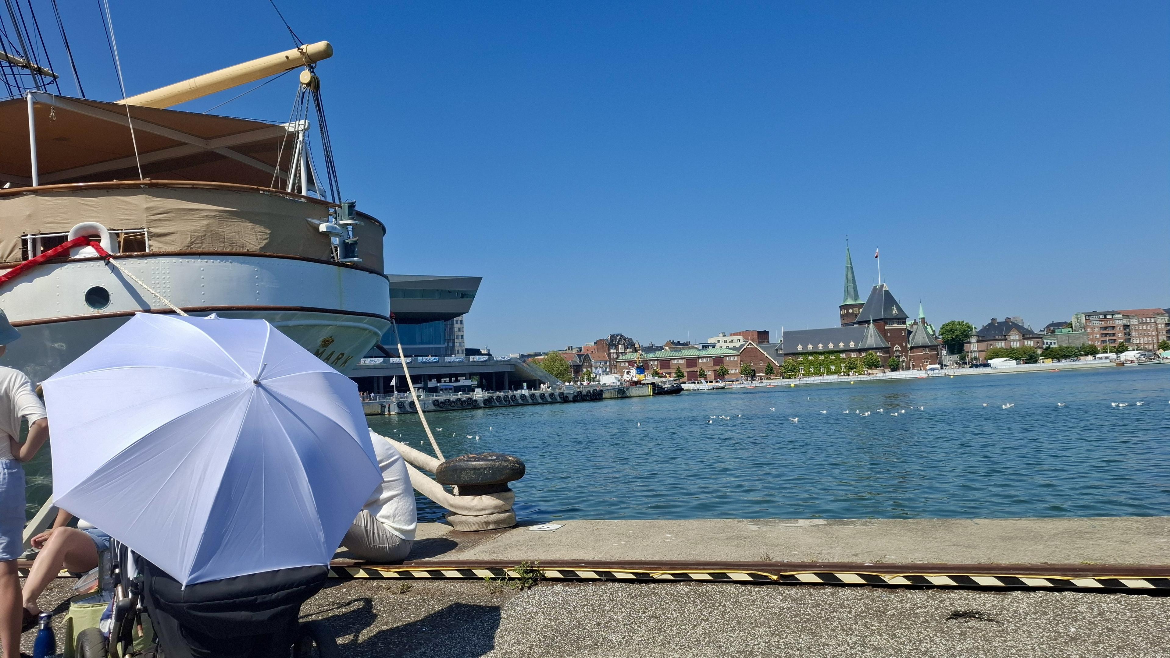 An open-tour ship moored at the harbour, blue sea and sky, Aarhus city skyline opposite. Just to the edge, a group (faces not shown) of people with an umbrella as a parasol shading children