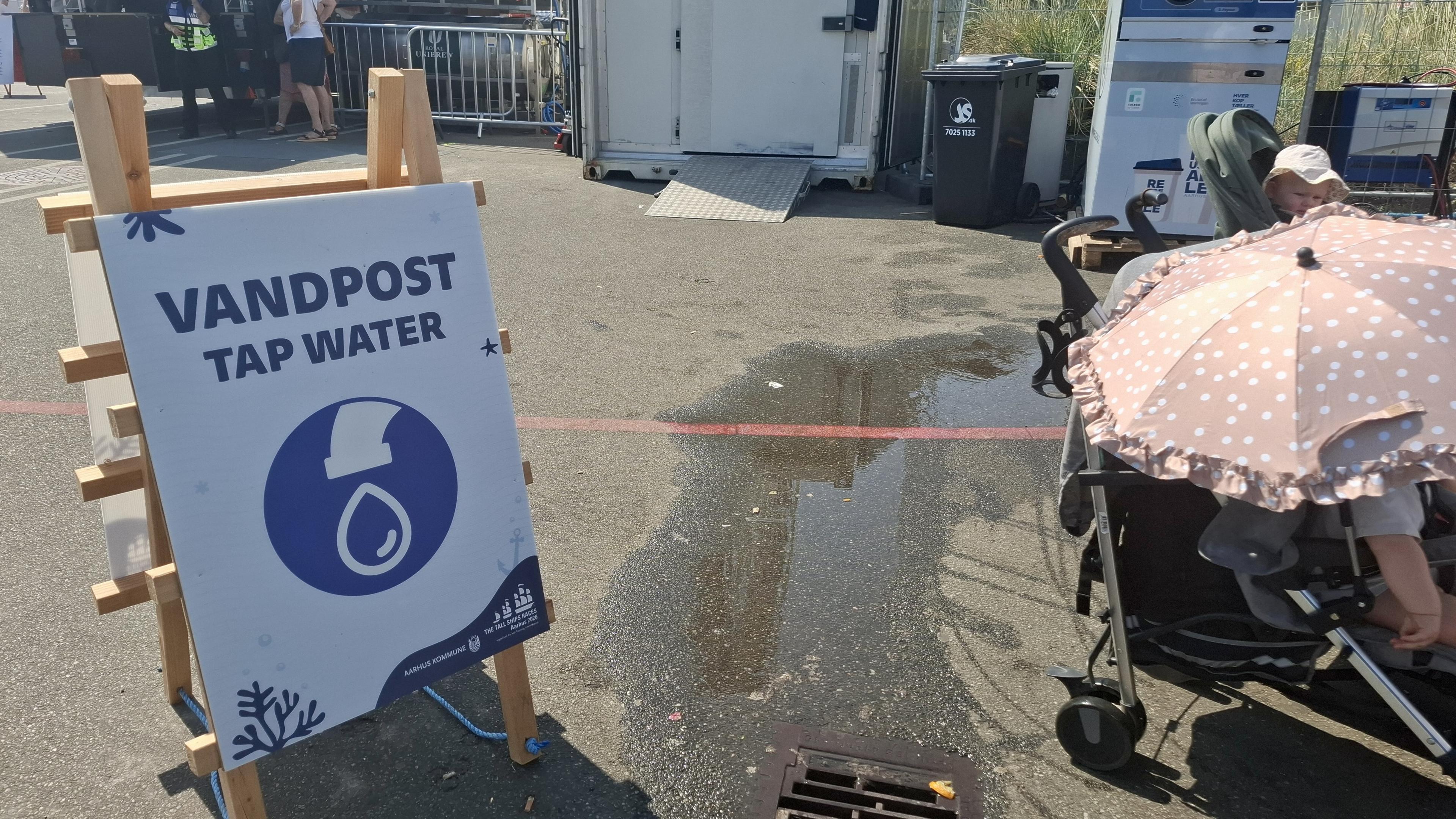 Sign for a tap water station at harbour. Beside it, the edge of a pram with a children's buggy with an umbrella giving it shade.