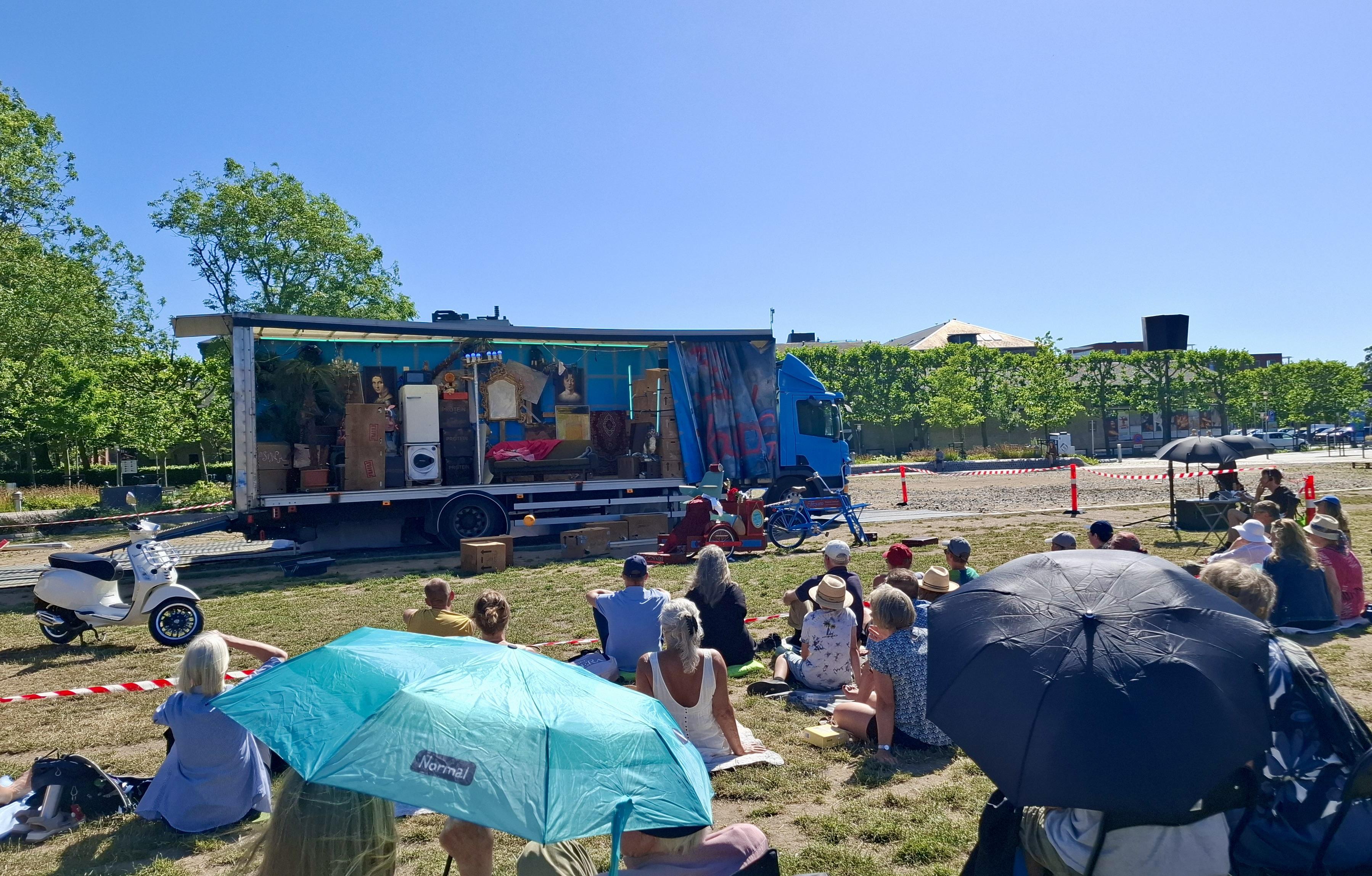 Short version: umbrellas as parasols in crowd. Fuller description: A small scattered crowd sitting on drought-browning grass in front of a stage. This is Opera on a Lorry, the stage is an actual lorry in a park in mid-city Aarhus. This is The Barber of Seville by Den Jyske Opera who are doing free Summer performances in nationwide parks, like wandering players. The heatwave hit them and audiences. In this image, we can see a black umbrella and a green one. The green one has the logo of the chainstore Normal on it.
