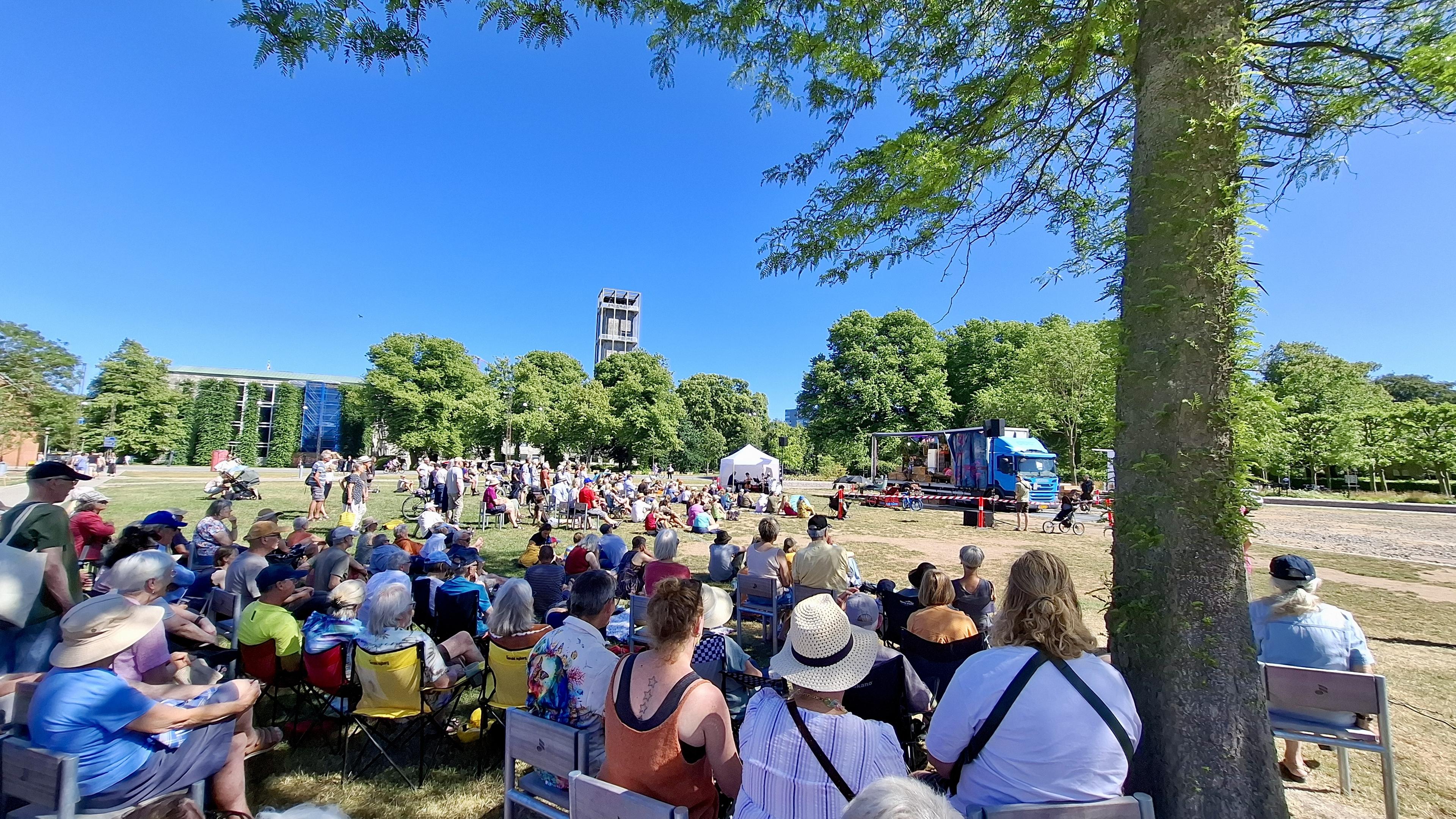 People crowded together under a single tree, far away from the stage of an outdoor event. The landmark Aarhus clock tower beyond under a cloudless blue sky. Fuller event description in the other photo's alt text.