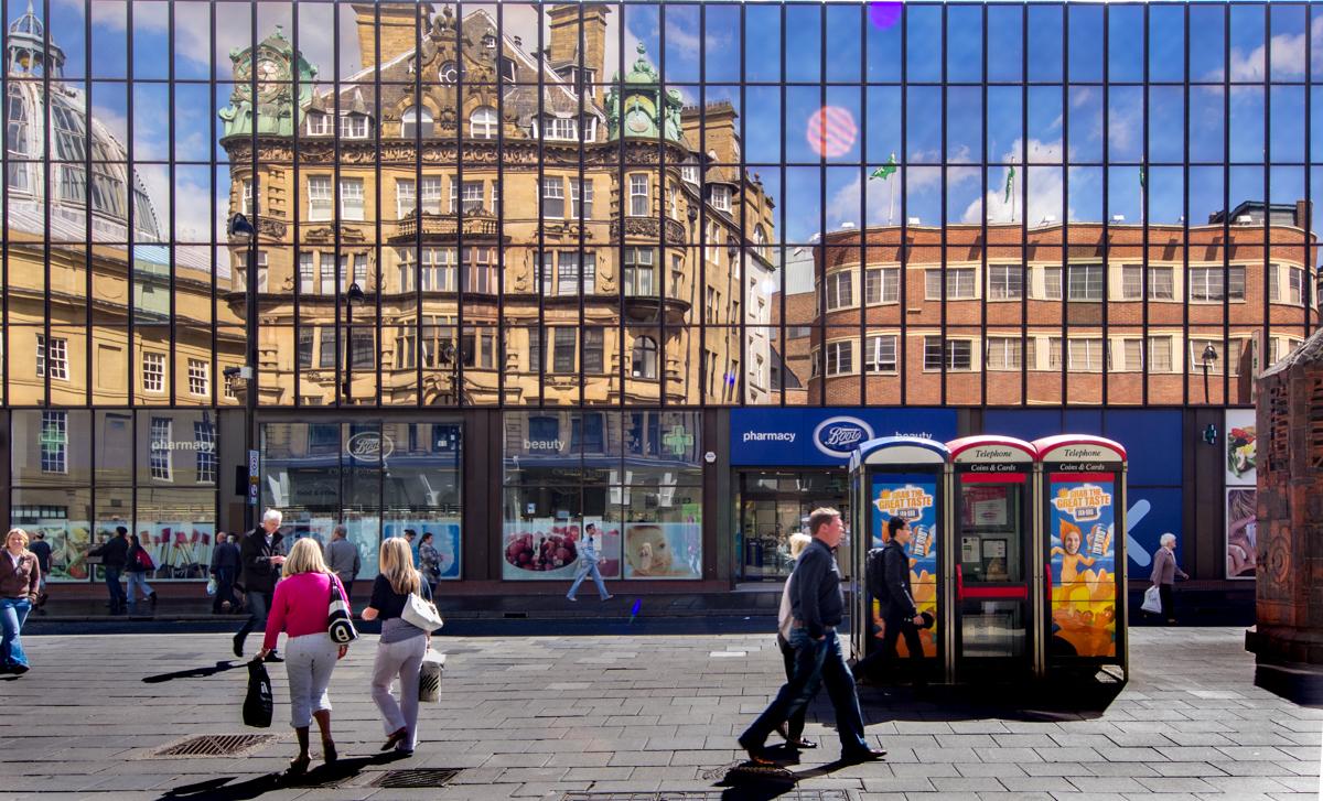 A modern city centre shopping street in the sunshine, the backdrop is a modern glass/windowed facade reflecting old buildings on the opposite side of the road.