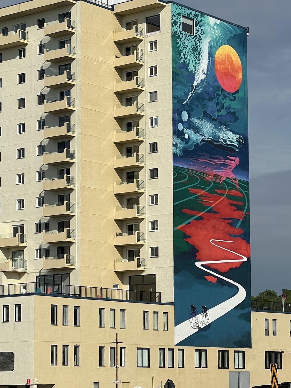 A beige brick apartment tower with many windows with a multicoloured mural of cyclists riding under a stylized space age sky. The base of the building has rows of tall narrow windows, while the upper portion shows small balconies and smaller windows on the left if the image and the mural in right centre and blue sky on the far right. The mural shows teal and bright red, with a white ribbon of road that the black silhouettes of cyclists ride. [Rimouski (QC, Canada) June 2026]