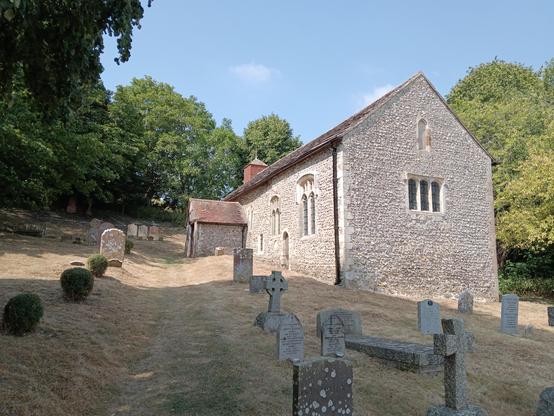 A single cell stone church set on sloping ground. A porch covers the entrance on the left (south) side. A number of randomly remaining gravestones standing in dry summer grass. Overhanging trees on the left side and in the background.