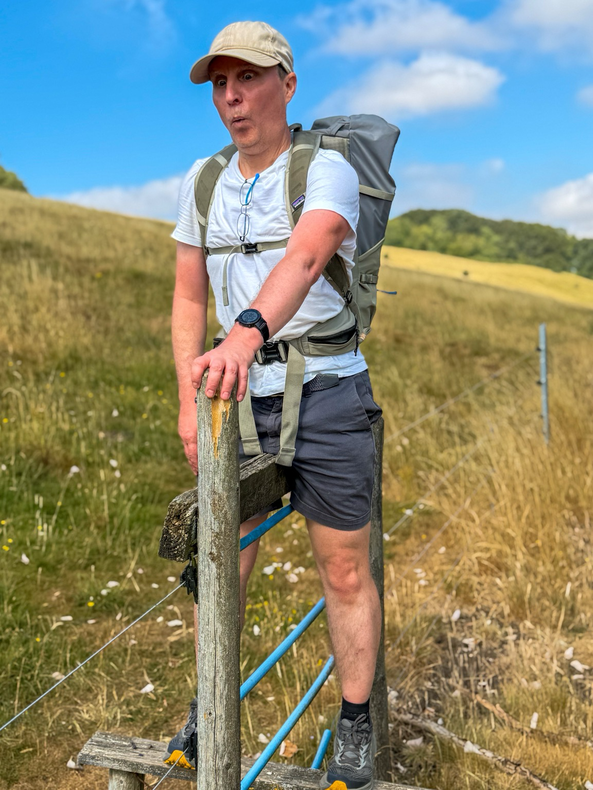 Me trying to get over a rather tall stile. I have both legs over but the wooden strut across the top of the stile is so high it’s crowding my underregions. I am holding a vertical stake and pulling a comical surprised pained expression. The background is a grassy meadow. I am wearing a white t-shirt, grey shorts, a beige cap and a large rucksack